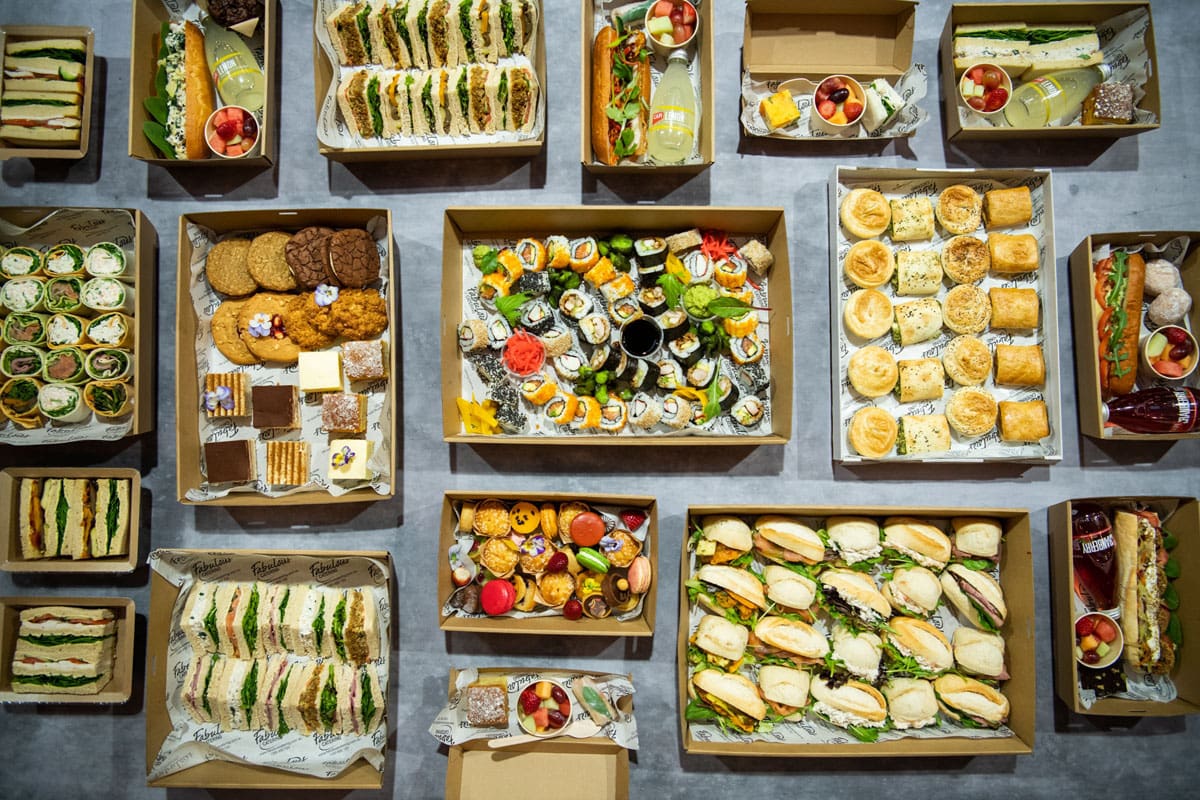 Overhead view of boxed catering platters: sandwiches, sushi, wraps, pastries and sweets